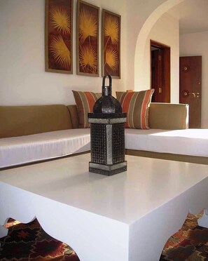 Una habitación con una mesa de centro blanca, un farol decorativo, un sofá con cojines y una puerta al fondo.