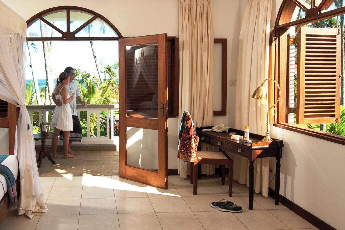 A hotel room with a bed, a desk, a chair, a window with a view of palm trees, and a door leading to an outdoor area.