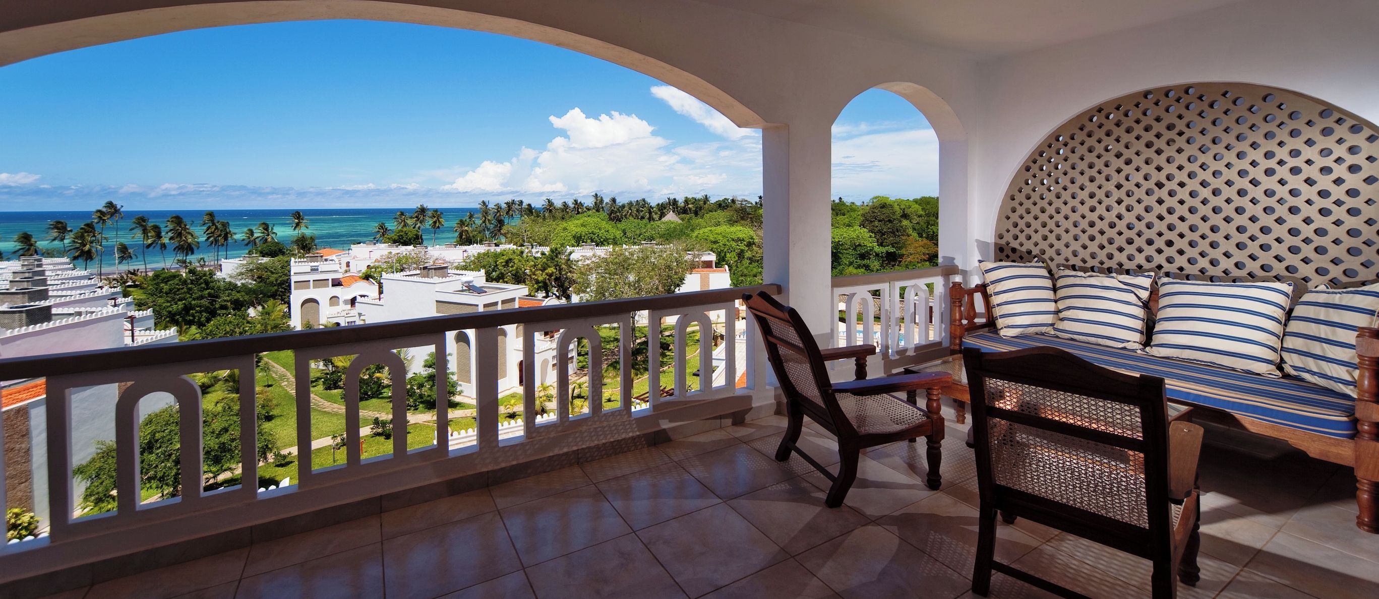 A balcony with a view of the ocean and buildings, a wooden chair, a bed with striped bedding, and a decorative headboard.
