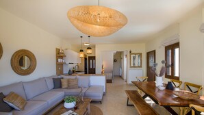 A spacious living area with a large sofa, wooden coffee table, and a dining table with chairs. A woven light fixture hangs from the ceiling, and there are wicker wall decorations. - EcoHome Villa (Chania)