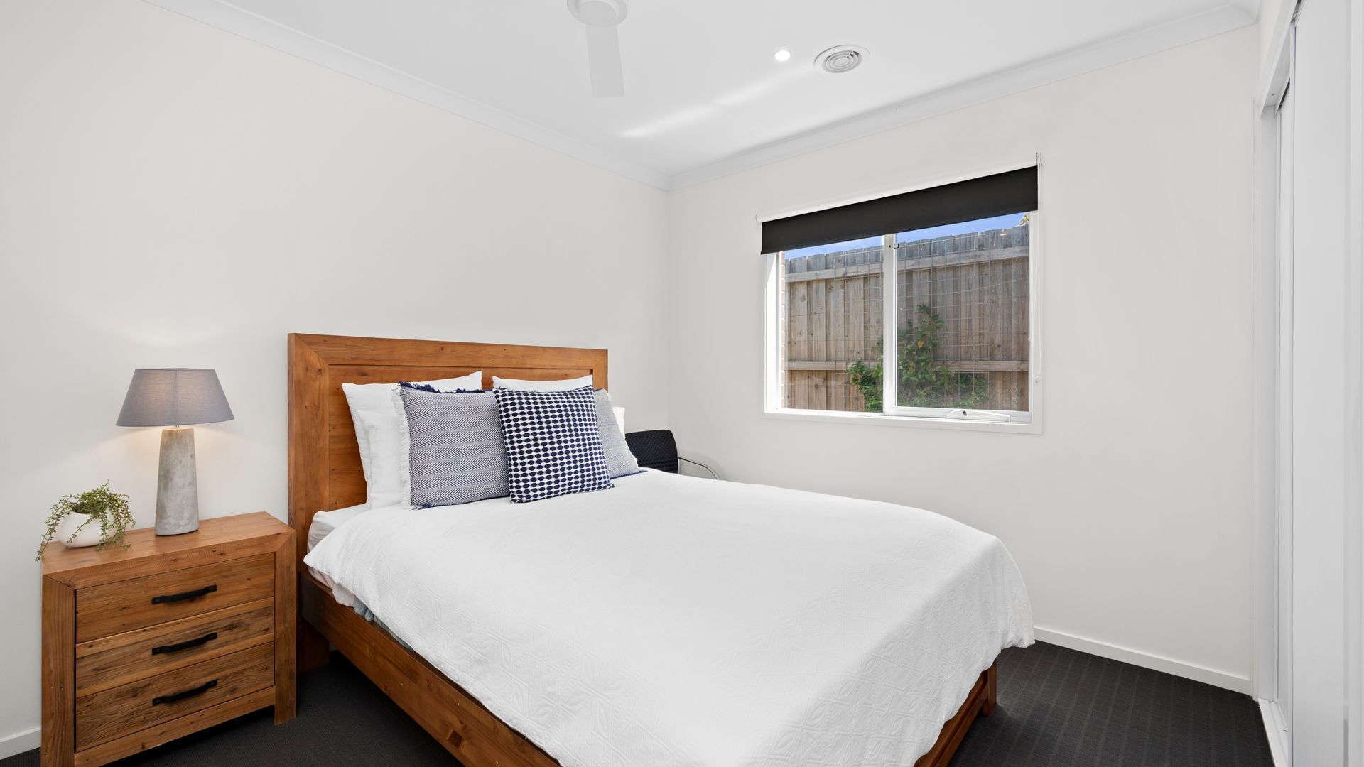 A bedroom with a wooden bed, a bedside table with a lamp, and a window with a view of a wooden fence.