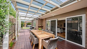 A covered patio with a wooden table and wicker chairs, a brick wall, and a glass door leading to an indoor area. - Aerial Beach House - Beach & Golf (Torquay)