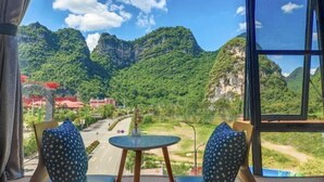 A room with a large window offering a view of a mountainous landscape. There is a small round table with a single chair in front of the window. - Yangshuo Xitian Yueshan Homestay (Eternal Love Store) (Yangshuo)