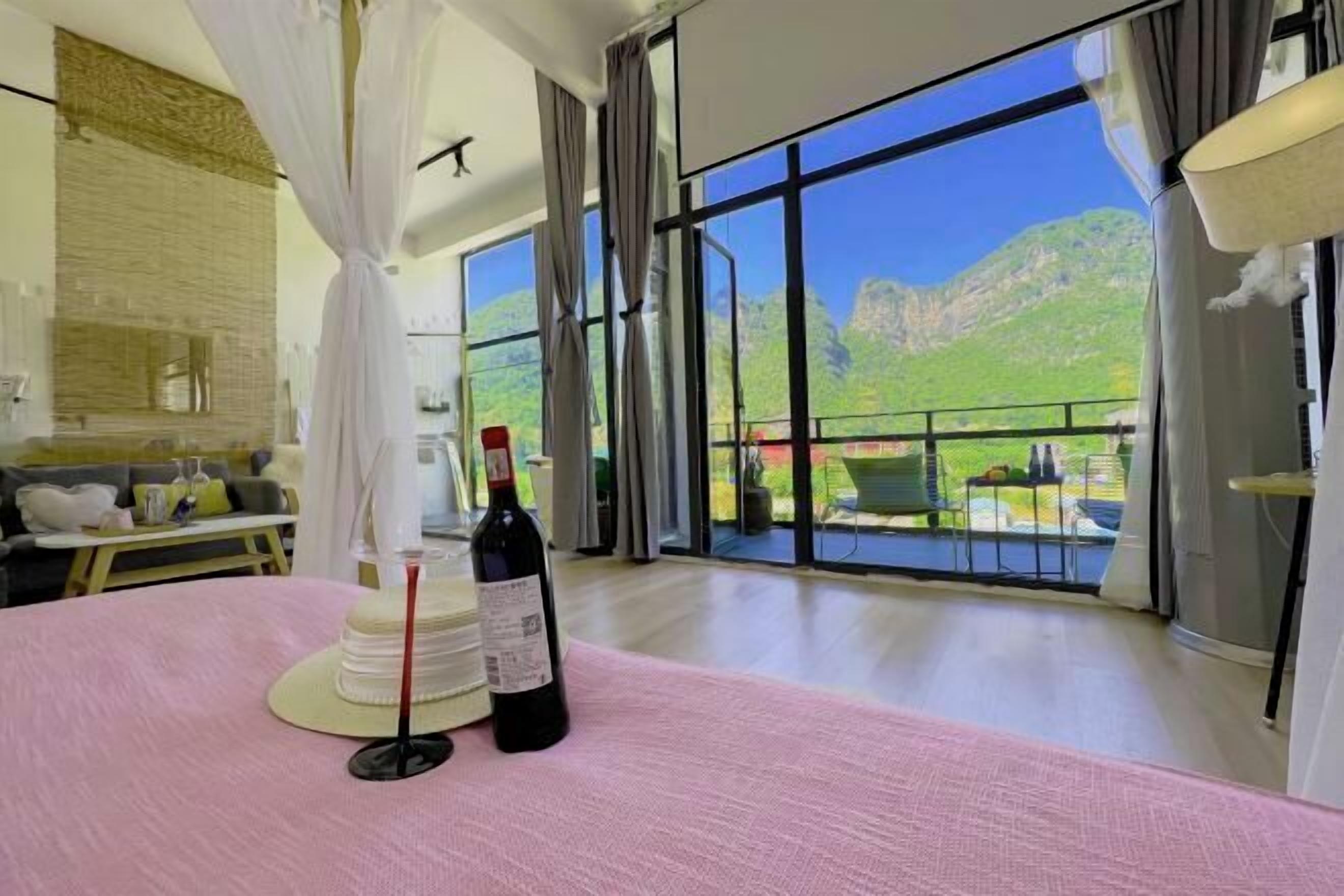Ein geräumiges Zimmer mit großem Fenster und Blick auf die Berge, einem Bett mit rosa Bettdecke, einer Weinflasche und einem kleinen Tisch mit Kerze und Glas.