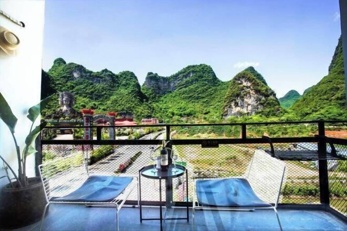 Un balcon avec deux fauteuils bleus dotés de coussins, une petite table ronde avec une bouteille et un verre, et une vue sur des montagnes verdoyantes.