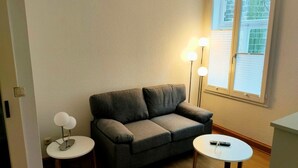 A small living room with a grey sofa, two round tables, and a window with blinds. - Apartment 1 Marktkirchhof - Hotel Theophano (Quedlinburg)