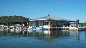 Un puerto deportivo con barcos amarrados, un edificio cubierto y un paisaje montañoso al fondo.