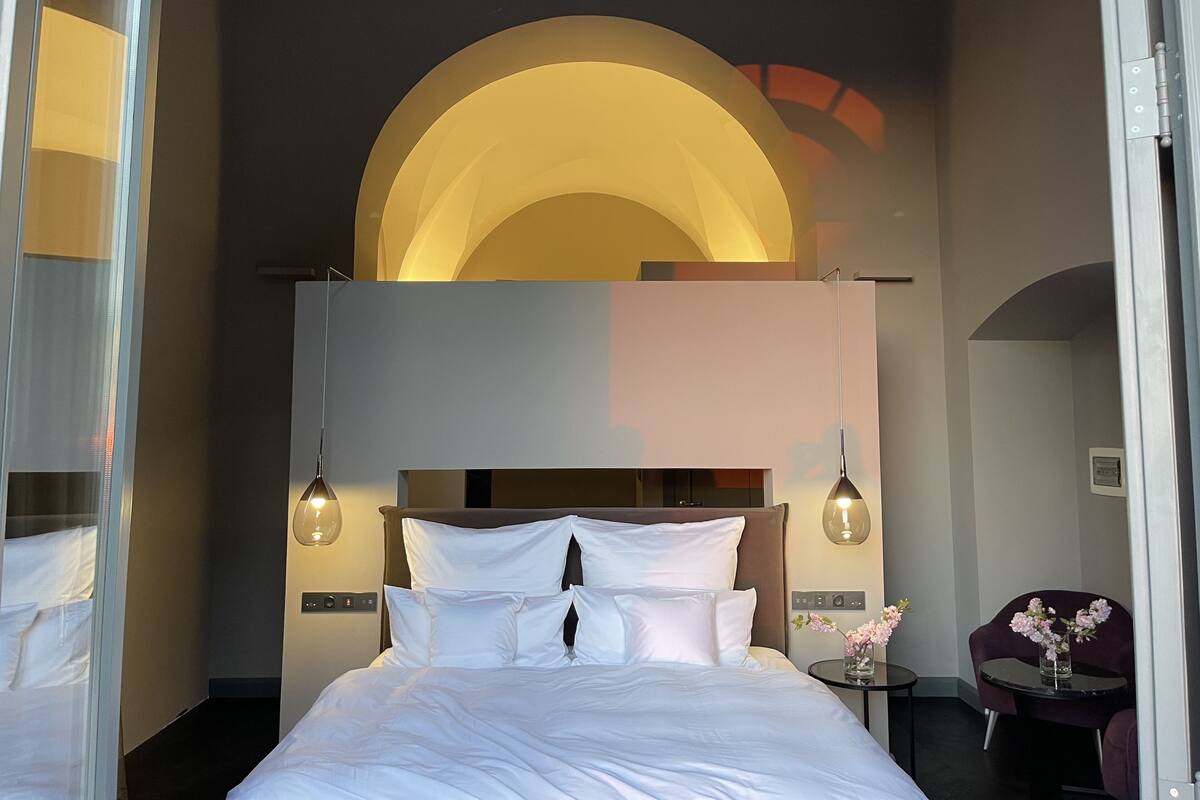 Una habitación de hotel moderna con una cama grande, dos lámparas colgantes, una pequeña mesa con un florero y vista a la ciudad a través de una ventana.
