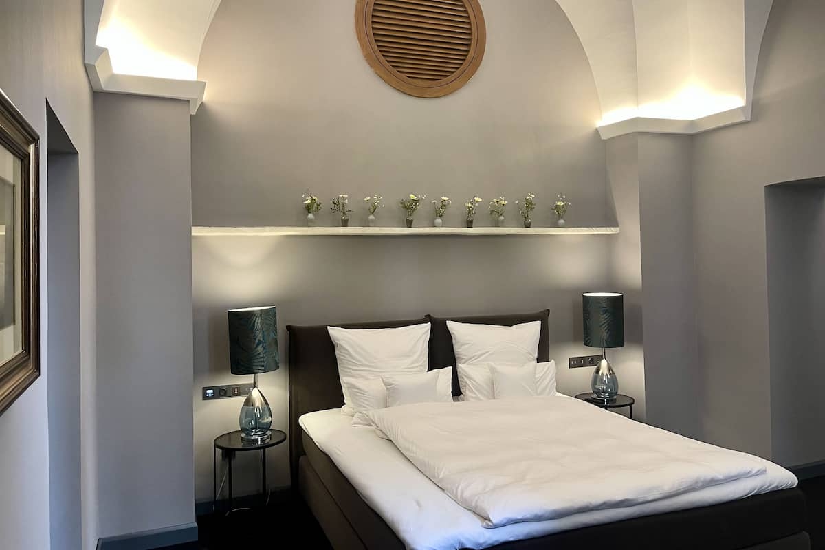 Habitación de hotel con cama, lámparas de noche, estante en la pared con flores y un respiradero circular en el techo.
