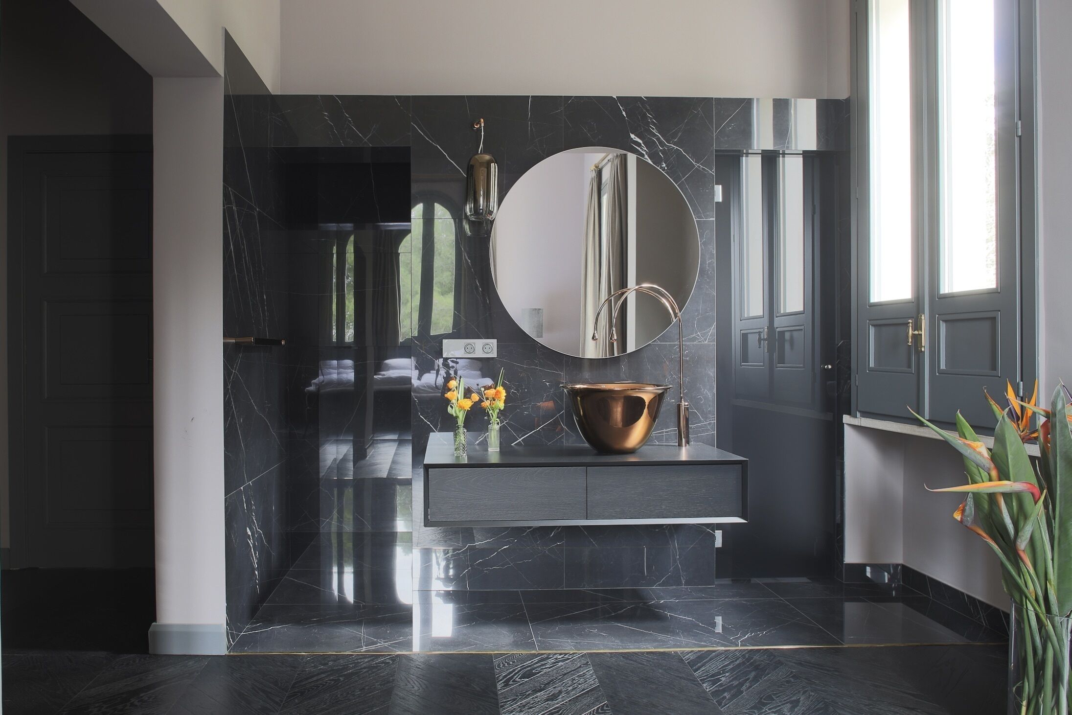 A modern bathroom with a dark marble countertop, a round mirror, a copper basin, and a vase with flowers.