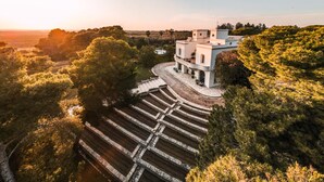 Un edificio grande y blanco con terraza y piscina, rodeado de árboles y colinas.