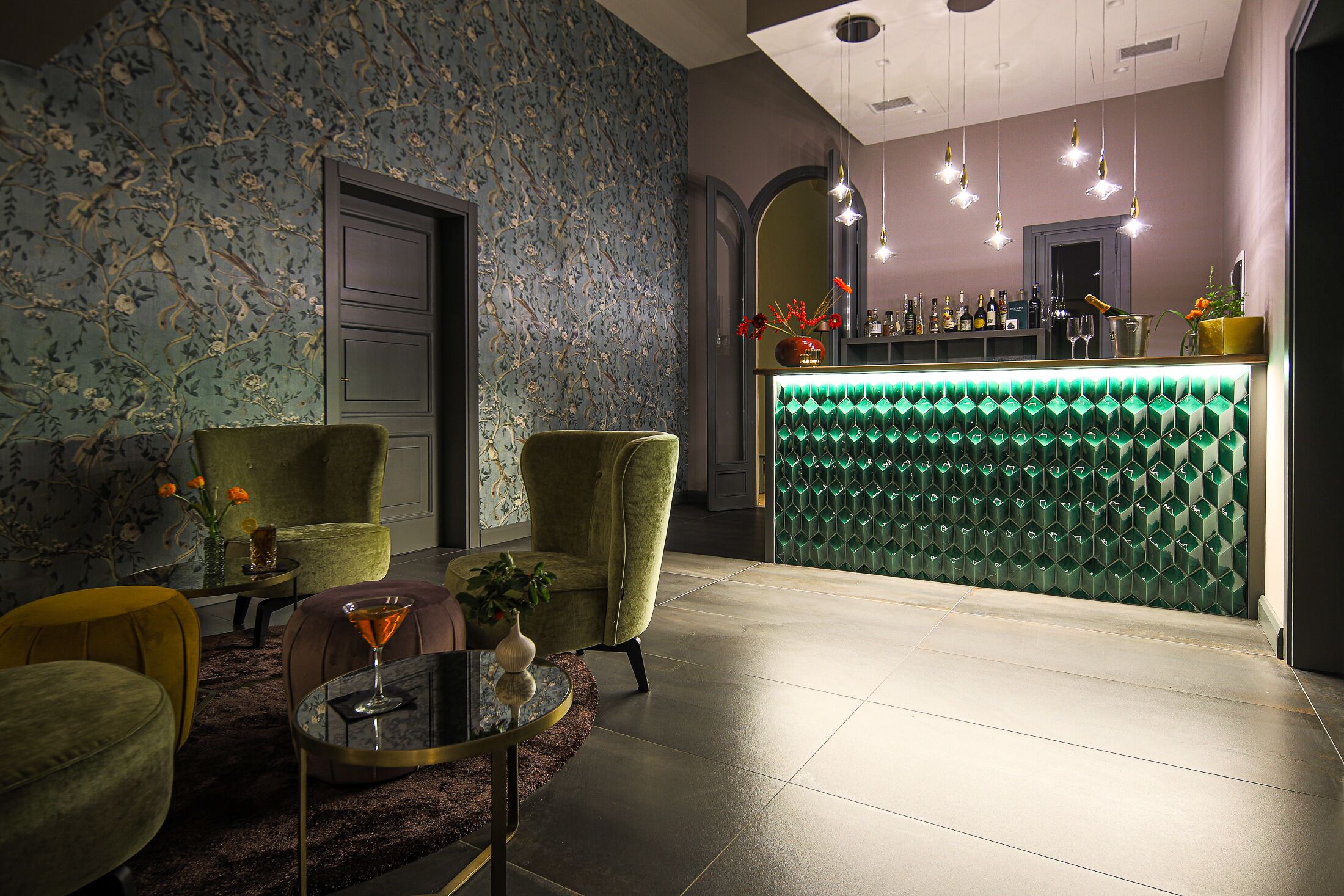 A bar area with a green neon-lit counter, a floral wallpapered wall, and a seating area with a round table and two armchairs.