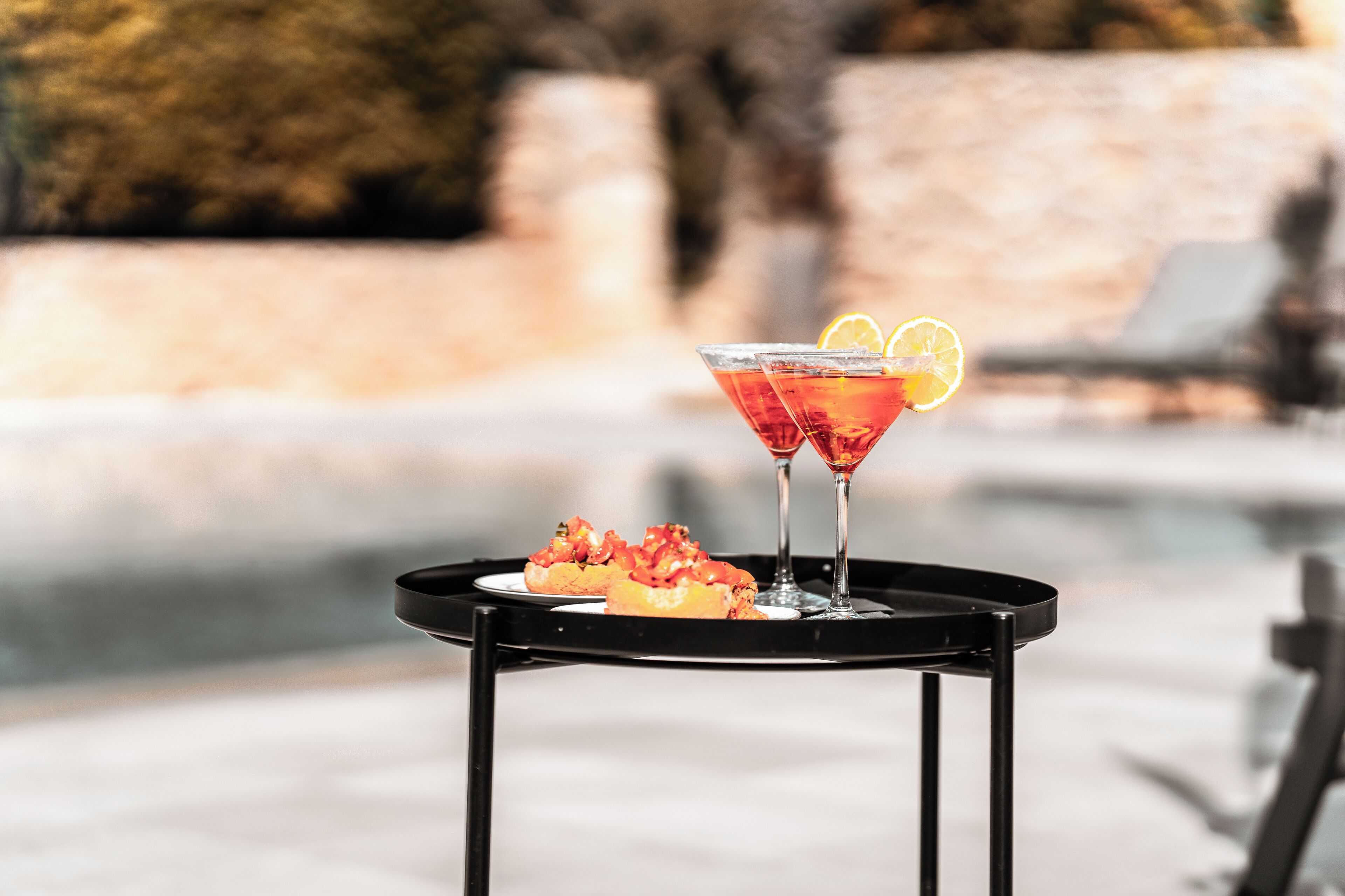 Un verre à martini, orné d’une tranche de citron, est posé sur un plateau noir, à côté d’une assiette remplie de nourriture, le tout étant placé sur une table au bord de la piscine.