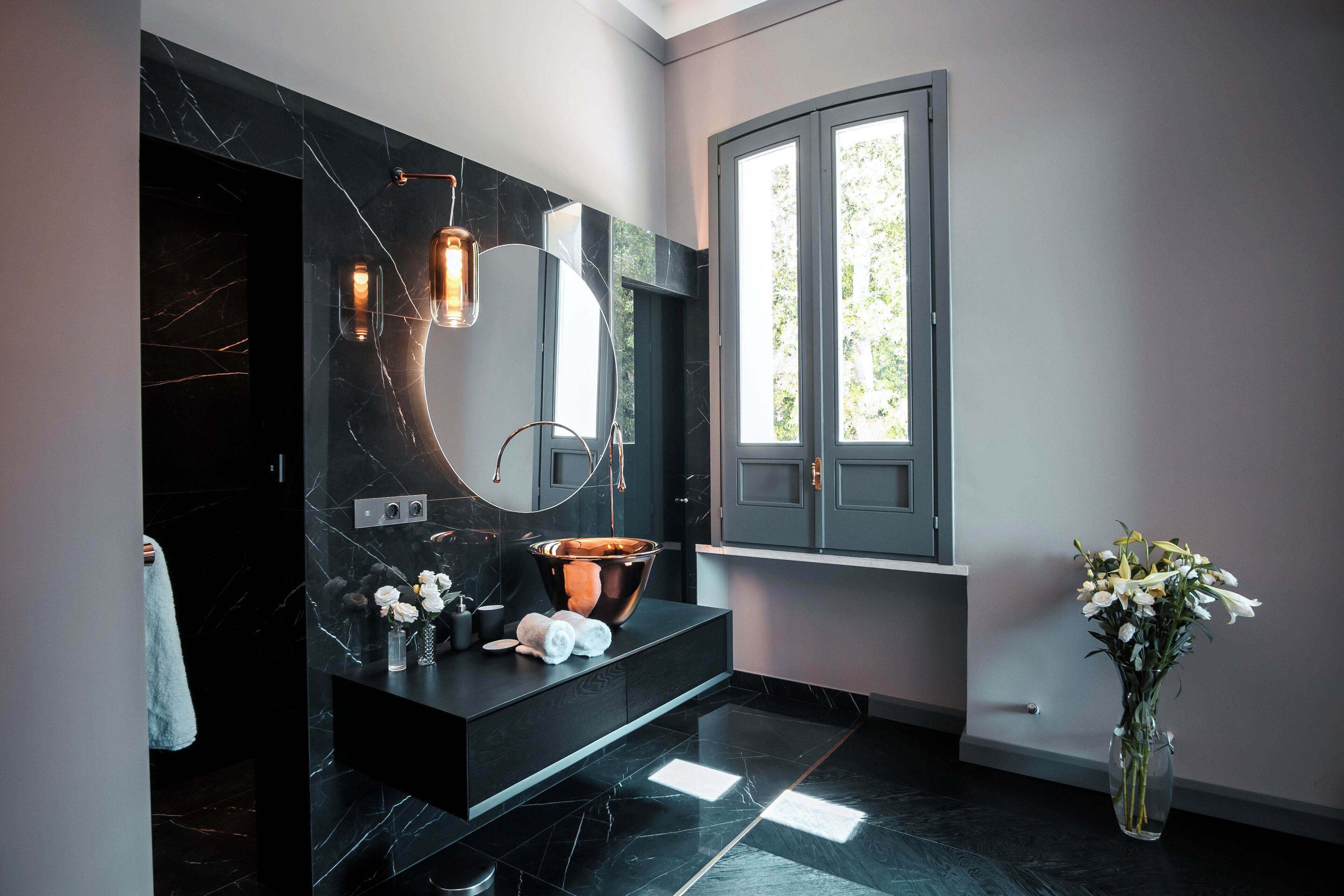 Un baño moderno con un tocador de mármol negro, un espejo grande y una ventana con vistas a árboles.
