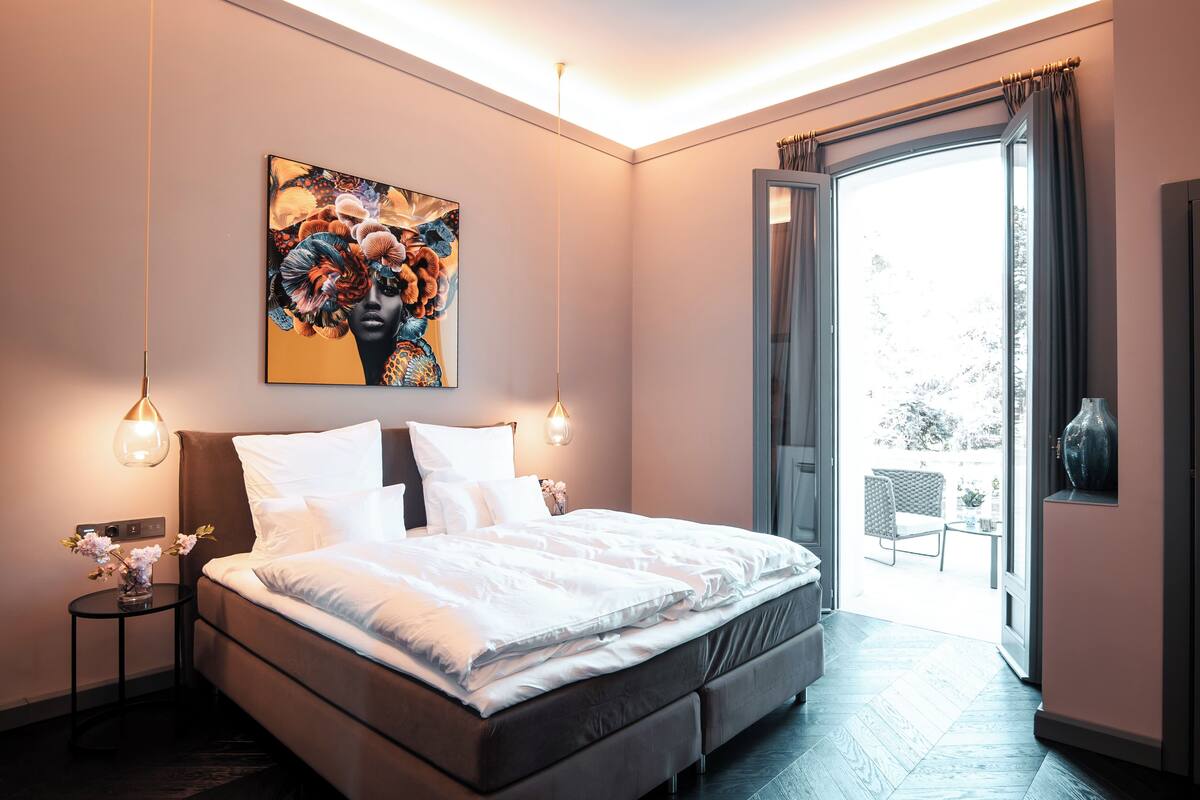 Un dormitorio moderno con una cama grande, un cuadro sobre la cama, una mesita de noche con un florero y vistas a través de una puerta abierta.