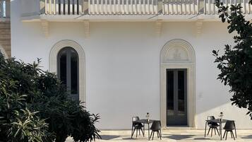 Un bâtiment à la façade blanche, doté de deux portes en arc de cercle et d’un balcon avec une balustrade. On y trouve quatre tables et des chaises disposées à l’extérieur.