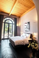 Un dormitorio con una cama grande, techo de madera, ventana con cortinas, un cuadro colgado en la pared y una mesita de noche con un jarrón de flores.