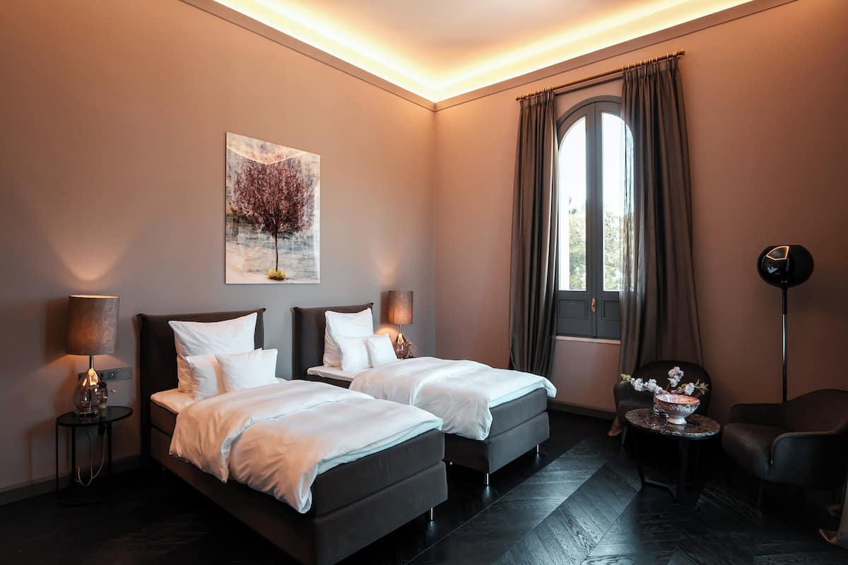 Habitación de hotel con dos camas, un cuadro, una ventana con cortinas, una silla, una mesa con un florero y una lámpara de pie.