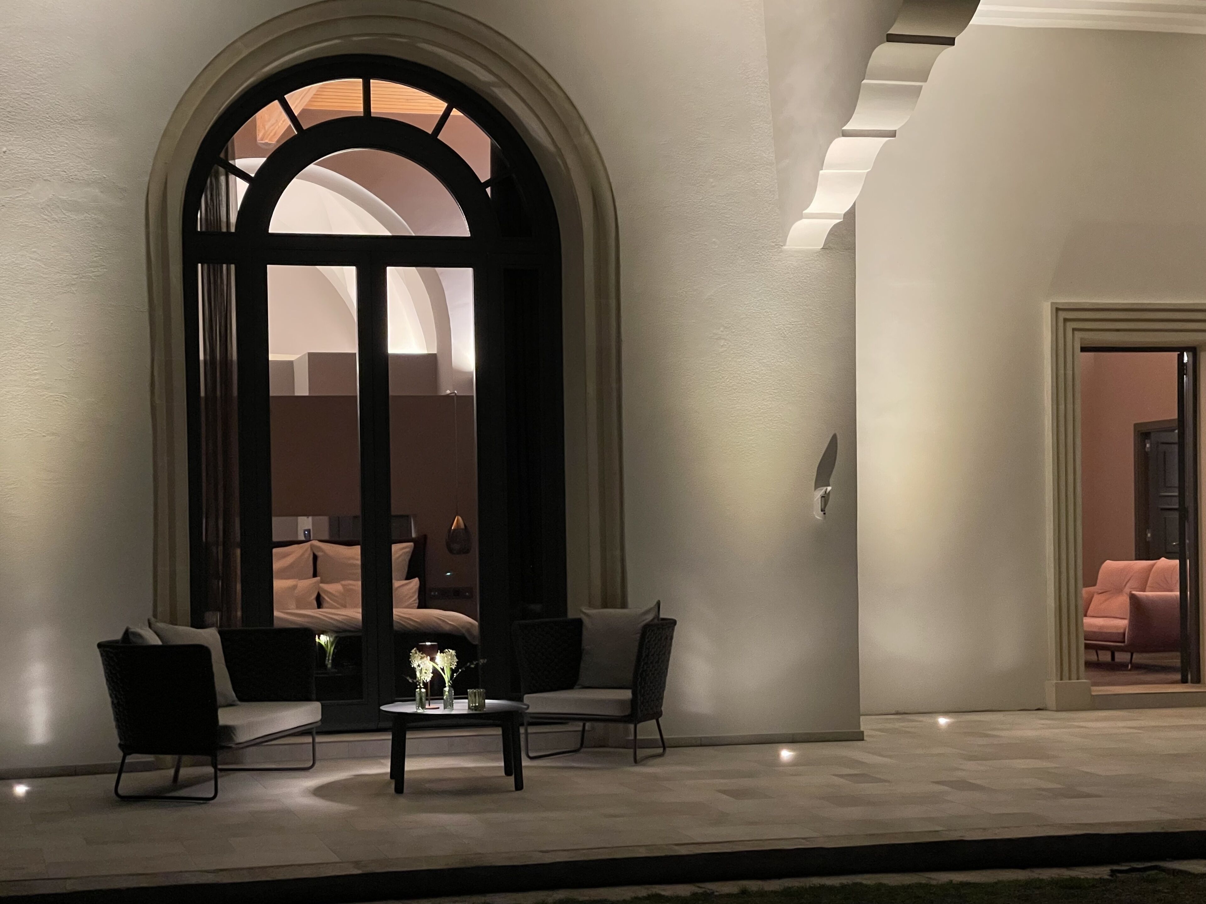 Un espace extérieur aménagé avec une table et des chaises, une grande porte en arc de cercle et une vue sur une pièce où se trouve un lit.