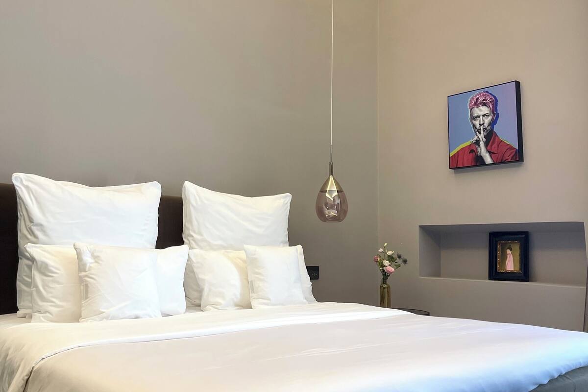 Un dormitorio moderno con una cama grande, ropa de cama blanca, una lámpara colgante y un cuadro en la pared.