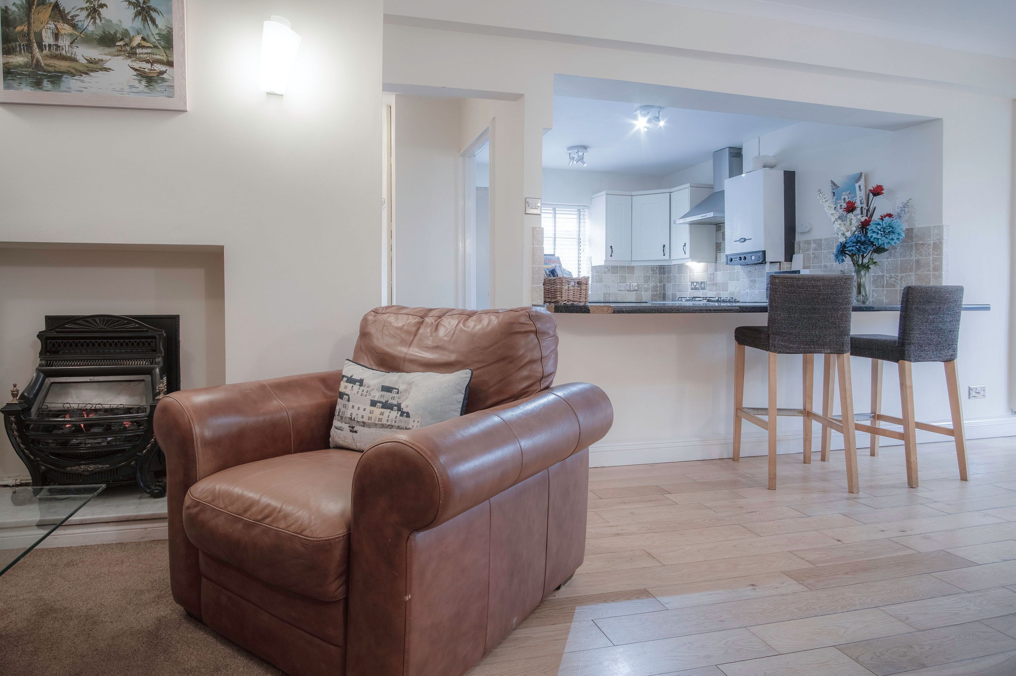 A living room with a brown leather sofa, a glass coffee table, a fireplace, and a kitchen visible through an open doorway.