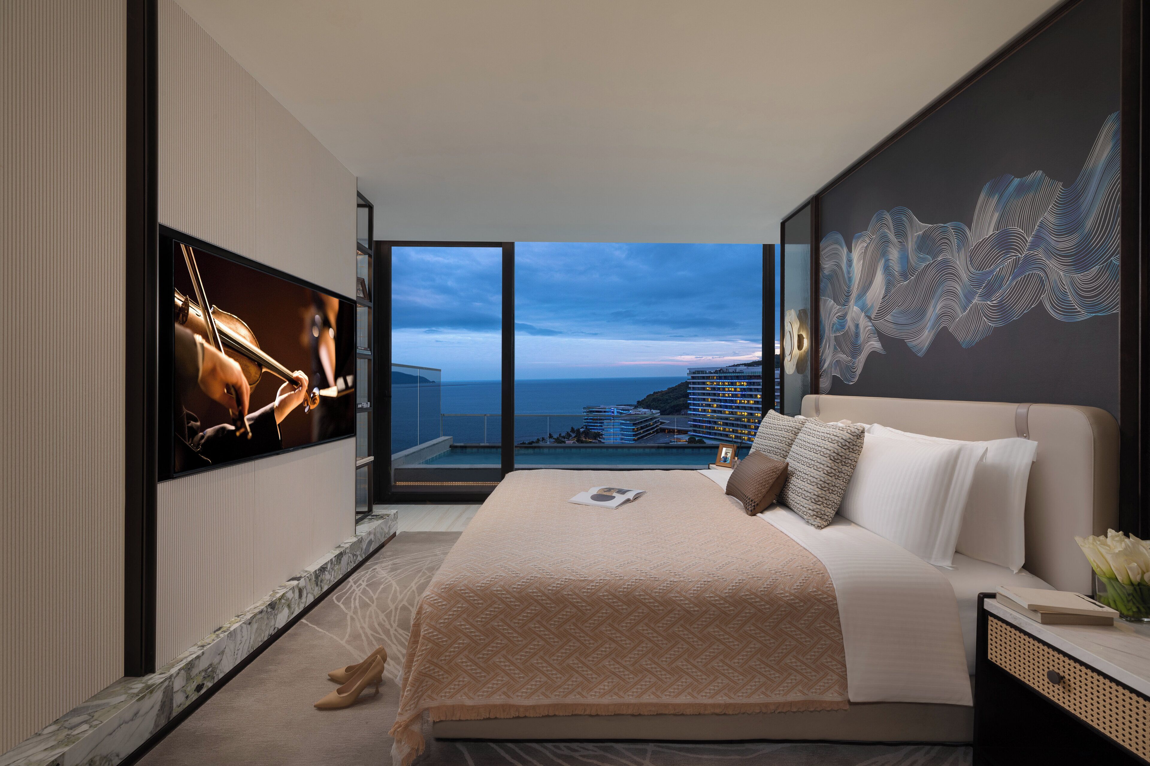 Un dormitorio moderno con una cama grande, un televisor en la pared y vistas al océano y edificios.