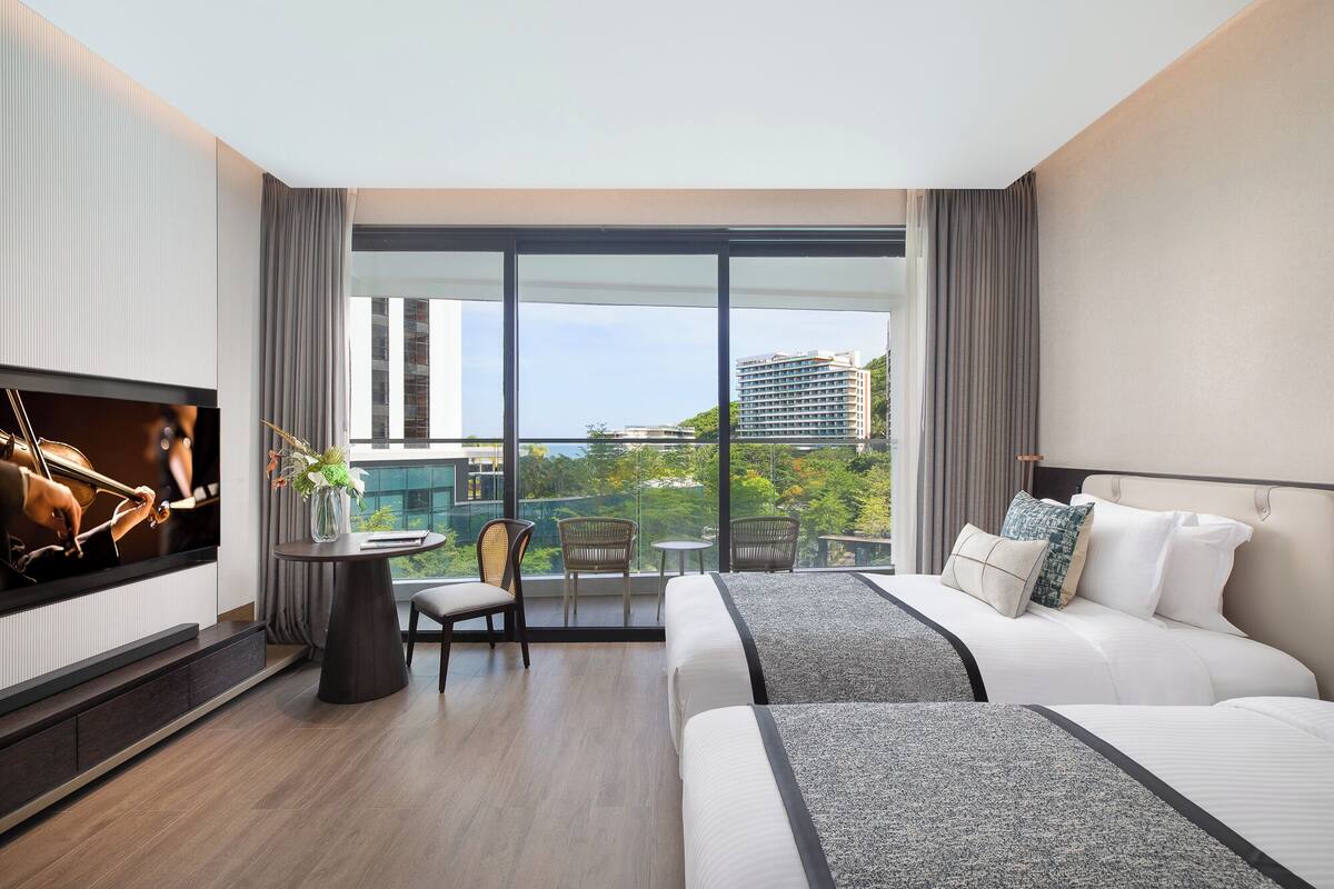 Ein modernes Hotelzimmer mit einem großen Bett, einem an der Wand befestigten Fernseher, einem Balkon mit Blick auf die Stadt und einem kleinen Tisch mit Stühlen.