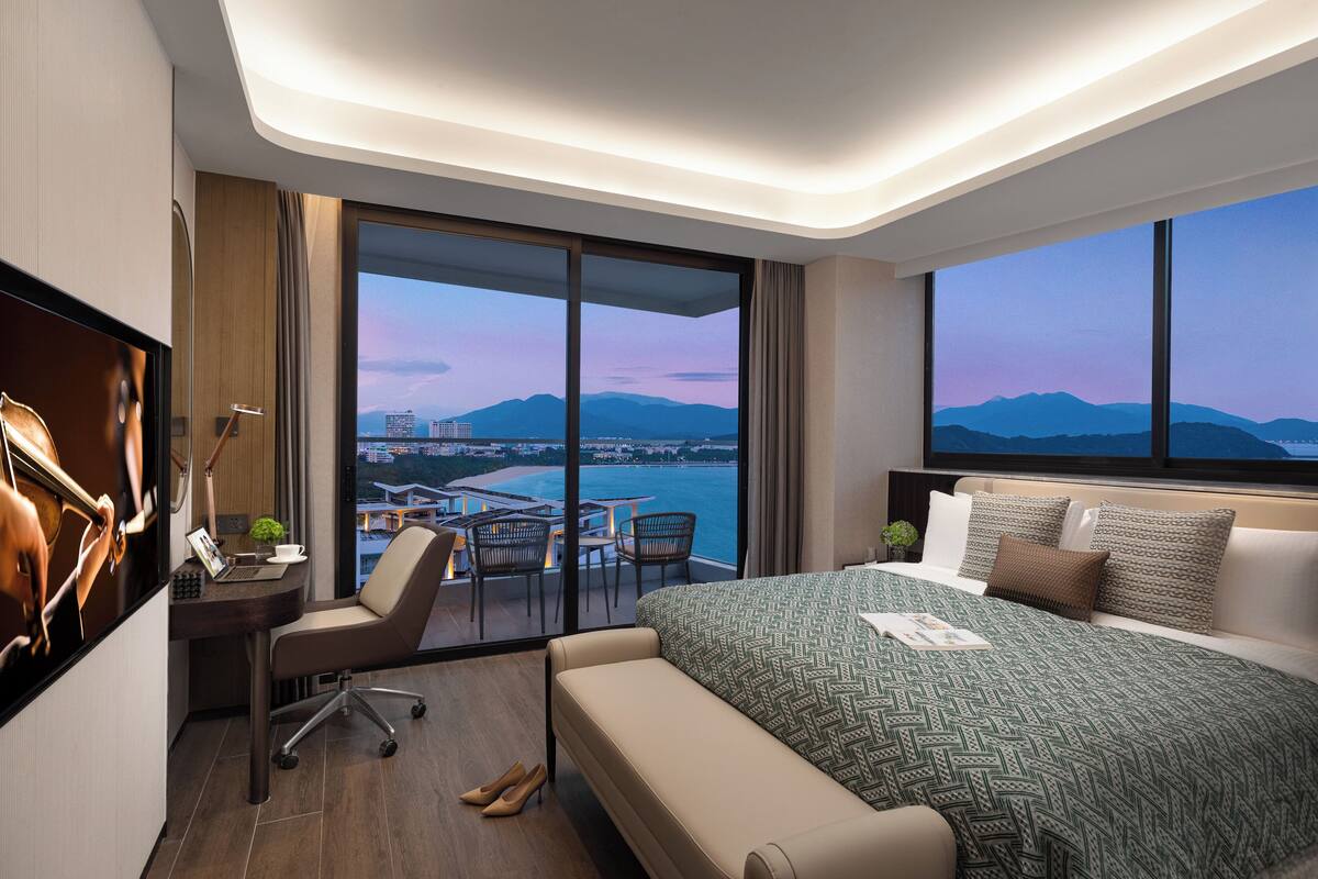 Ein modernes Hotelzimmer mit einem großen Bett, einem Schreibtisch mit einem Laptop, einem Stuhl und Blick auf Stadt und Meer.