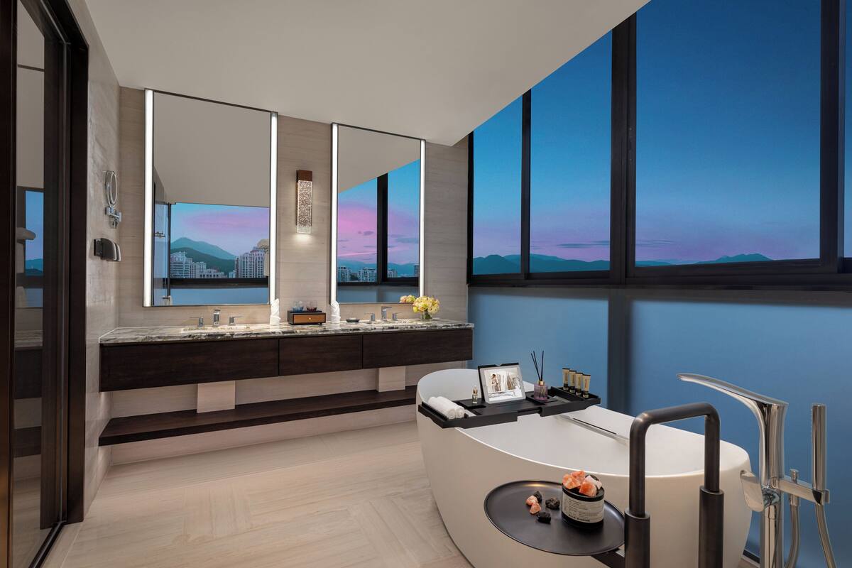 Ein modernes Badezimmer mit einer großen Badewanne, einem Waschtisch mit Spiegel und Blick auf die Stadt durch große Fenster.