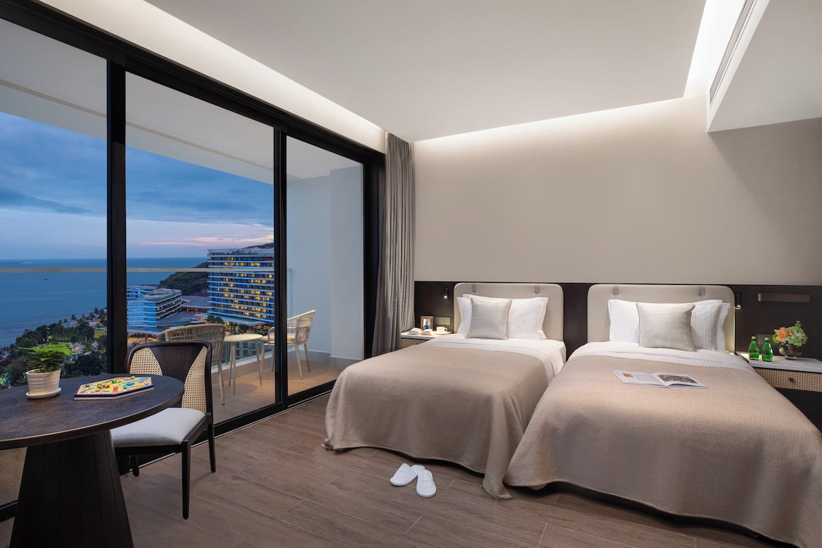 Ein Hotelzimmer mit zwei Betten, einem Balkon mit Blick auf das Meer und Gebäude, einem Tisch mit einem Stuhl und einem kleinen Tisch mit Pflanze und Buch.