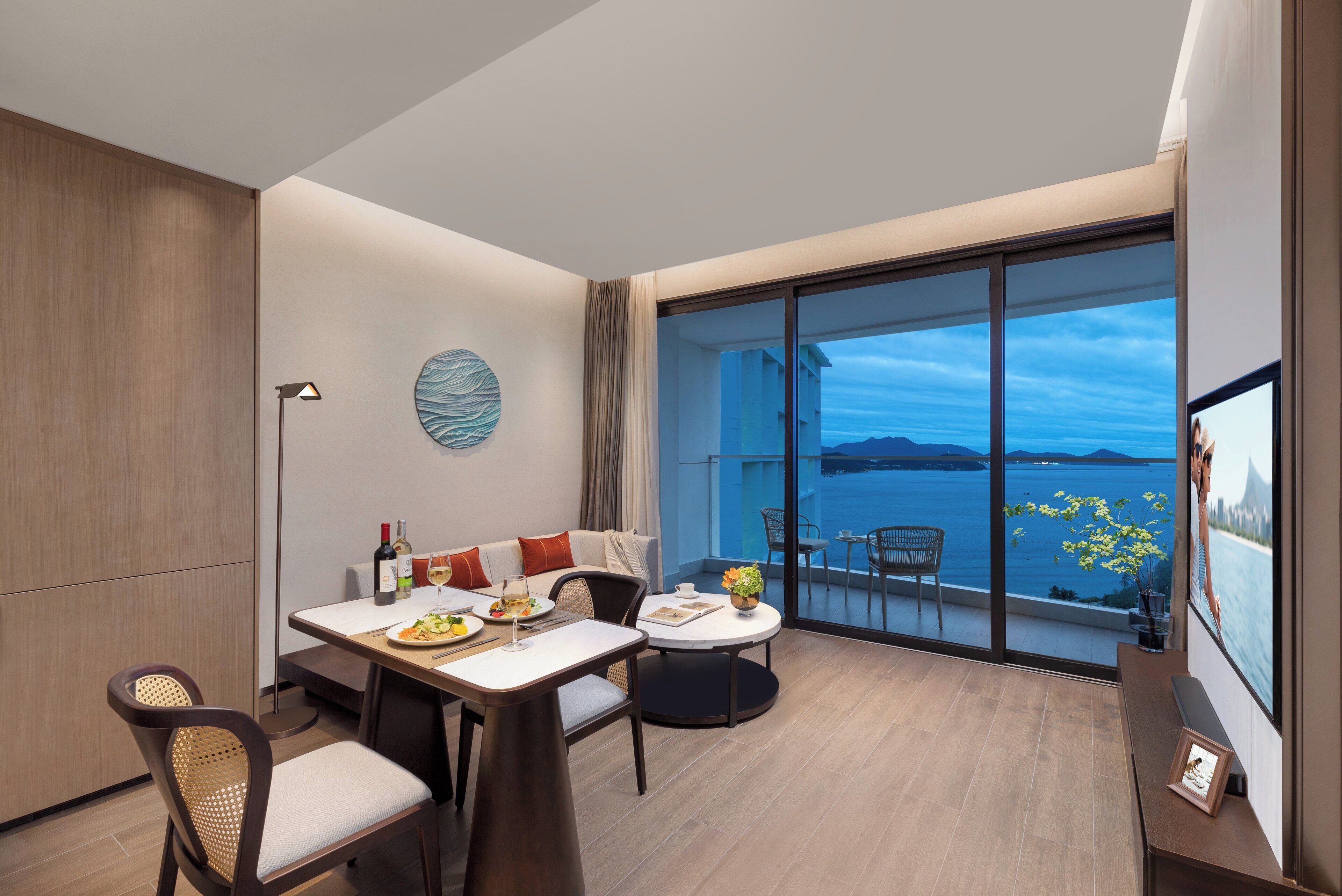 A modern room with a dining area, a sofa, a table with a vase of flowers, and a large window with a view of the sea.