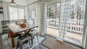 A dining area with a wooden table, chairs, and a bench. There are curtains on the windows, and a view of a deck and trees outside.