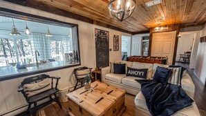 A cozy living room with a wooden ceiling, a large window, a wooden chest, a sofa with 'AHOY!' pillows, and a 'Laundry' sign on the door.