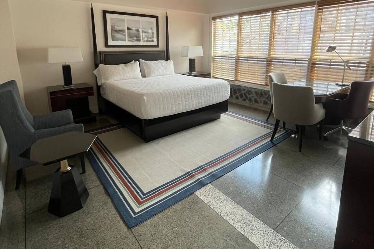 A modern hotel room with a bed, desk, chairs, and large windows.