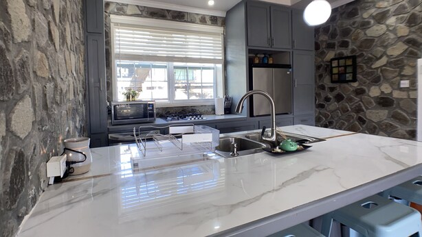 A modern kitchen with a stone accent wall, a large island with a sink, and a built-in oven. - Stonetrail (Villa)