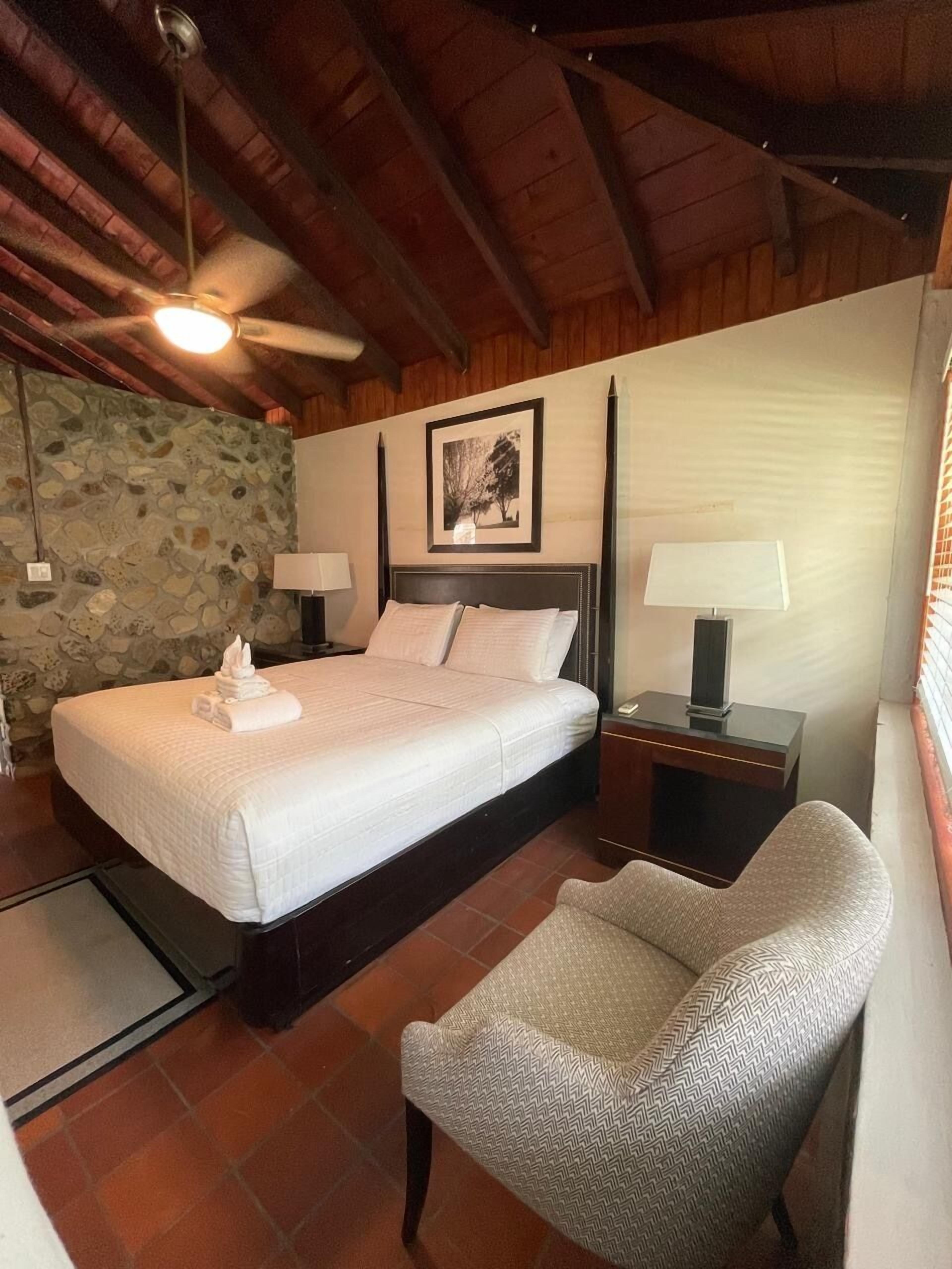 A bedroom with a bed, two bedside tables, a chair, a stone wall, and a ceiling fan.
