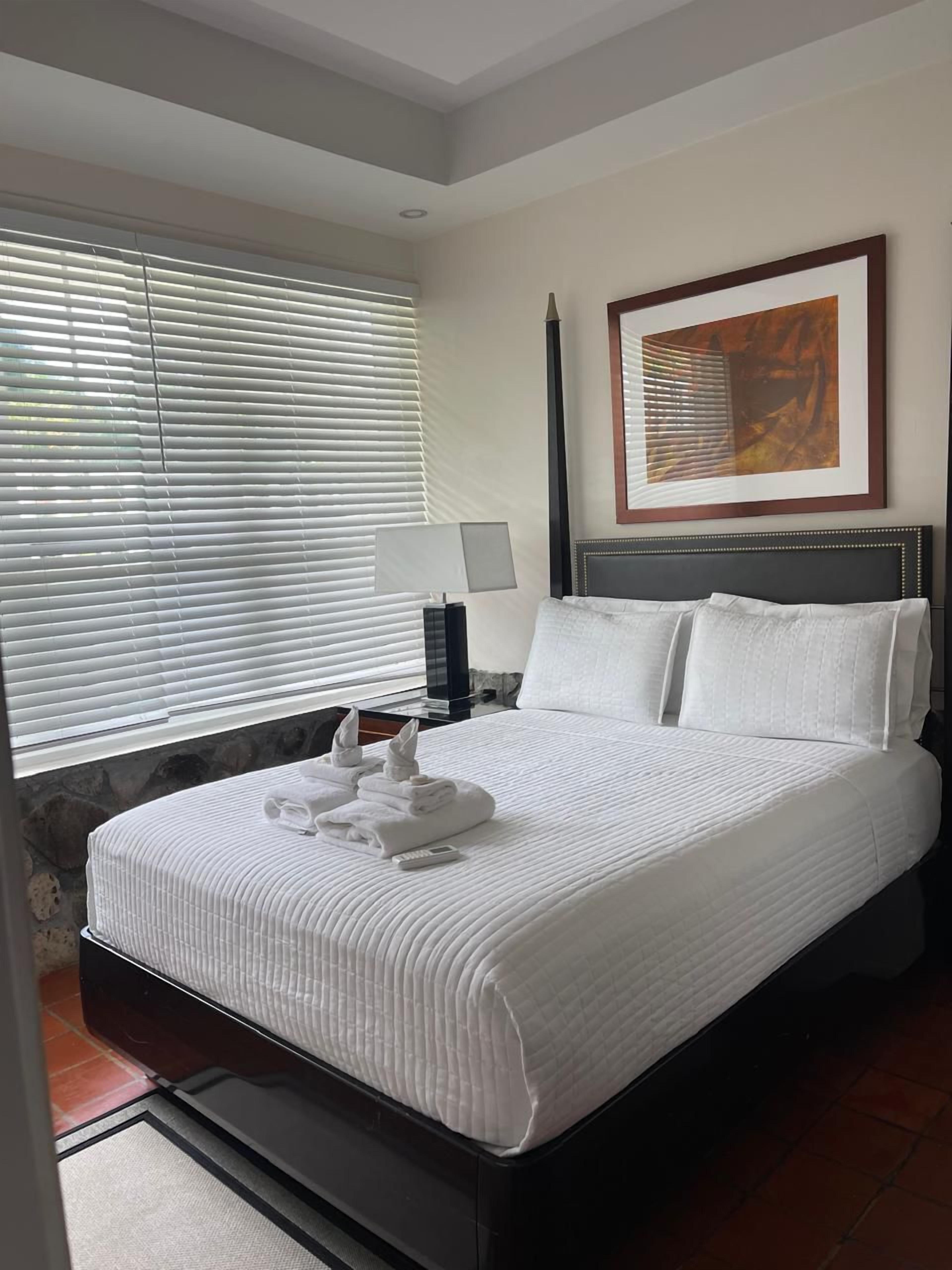A hotel room with a bed, a nightstand, a lamp, a framed picture on the wall, and a window with blinds.