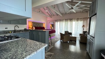 A modern kitchen with granite countertops, a gas stove, and a ceiling fan. There is a dining area with a table and chairs, and a living room with a sofa and a TV.