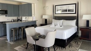 A hotel room with a bed, two bedside tables, a desk with a chair, a kitchen area, and a bar stool. - Stonetrail (Villa)