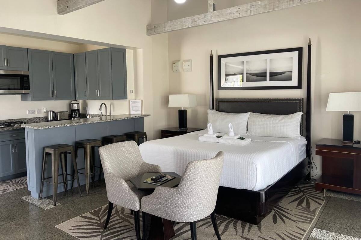 A hotel room with a bed, two bedside tables, a desk with a chair, a kitchen area, and a bar stool.