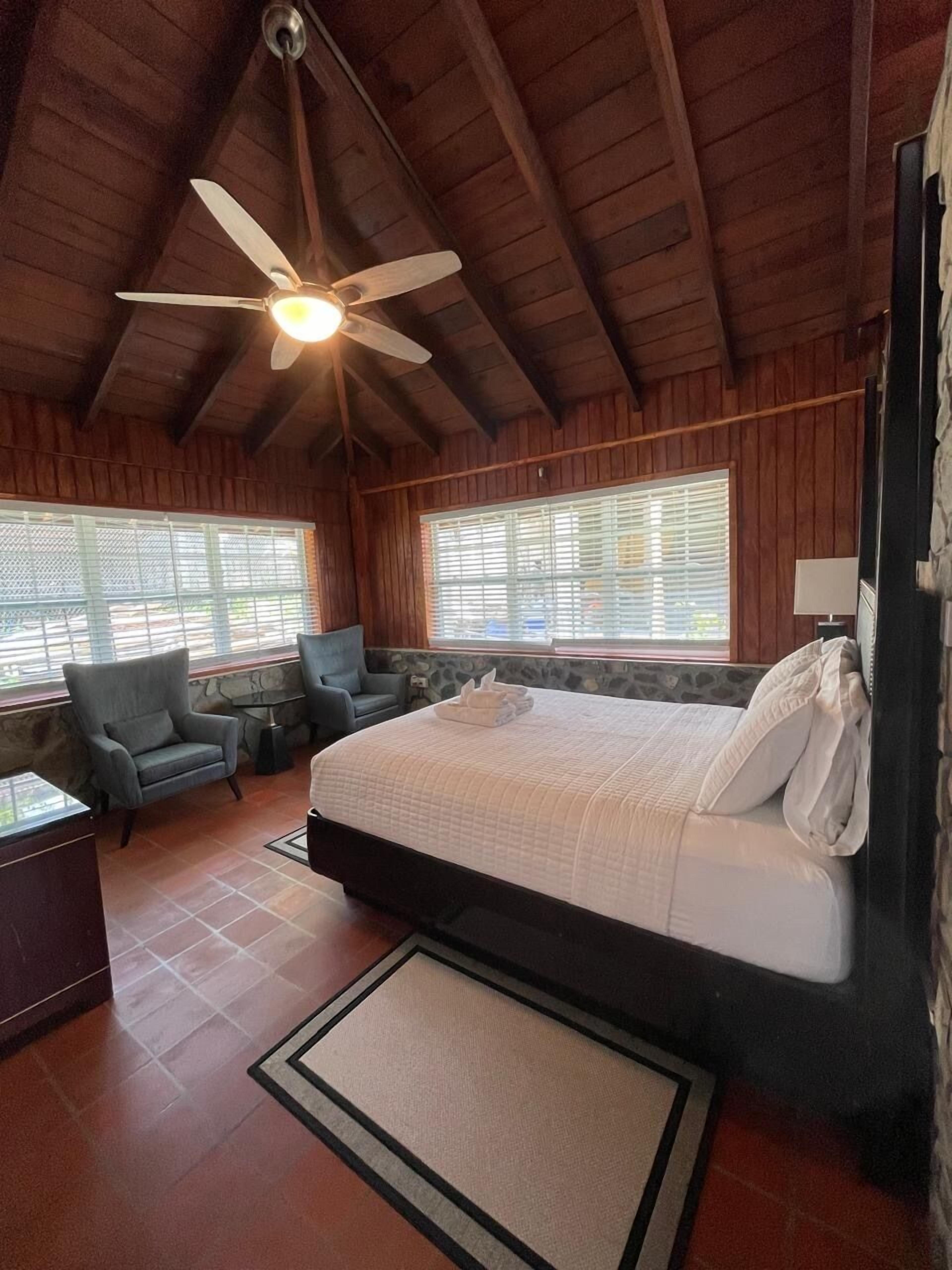 Un dormitorio de cabaña de madera con una cama, dos sillones, un ventilador de techo y ventanas amplias.