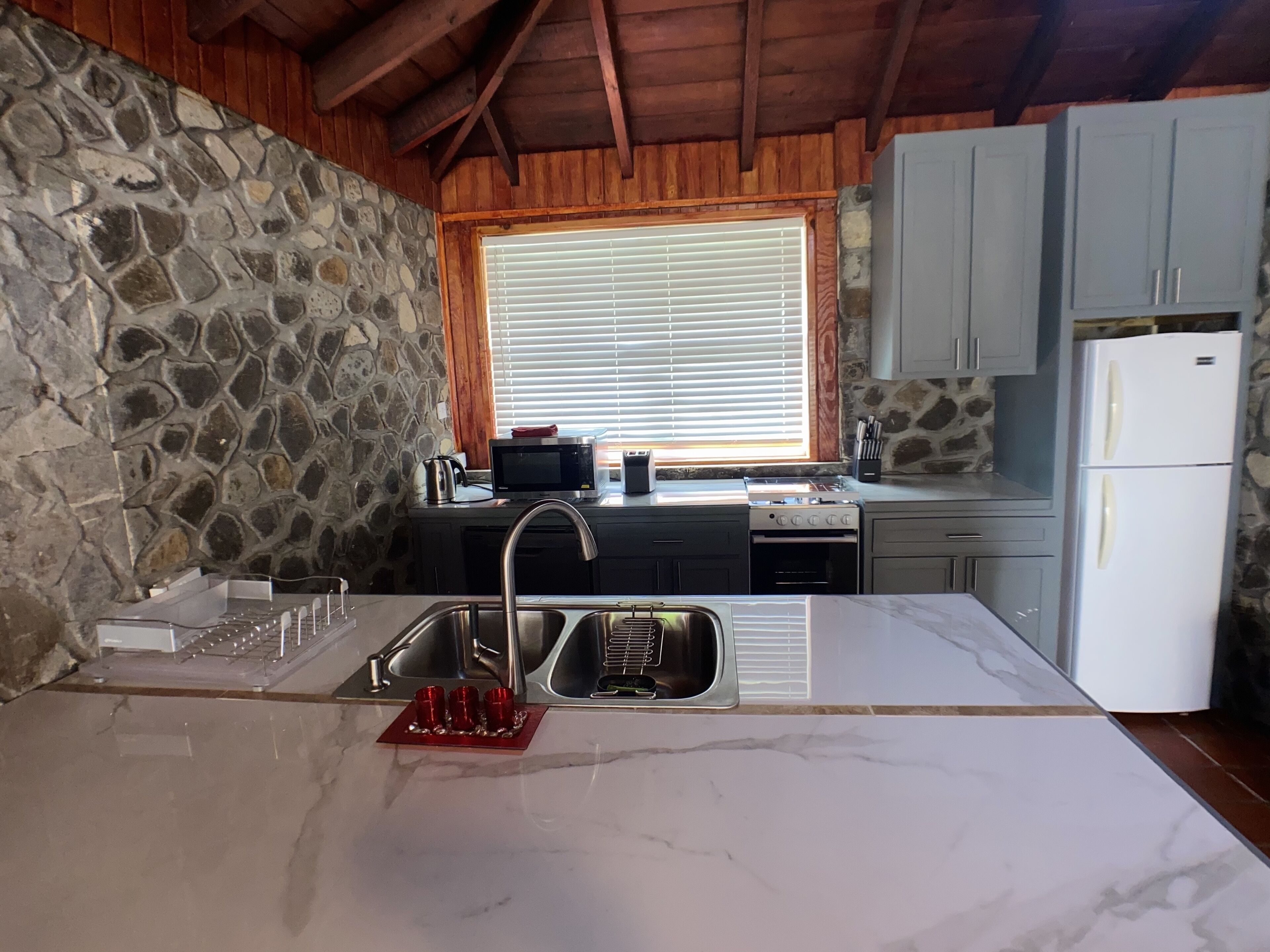 A kitchen with a stone wall, wooden ceiling, and a window with blinds.