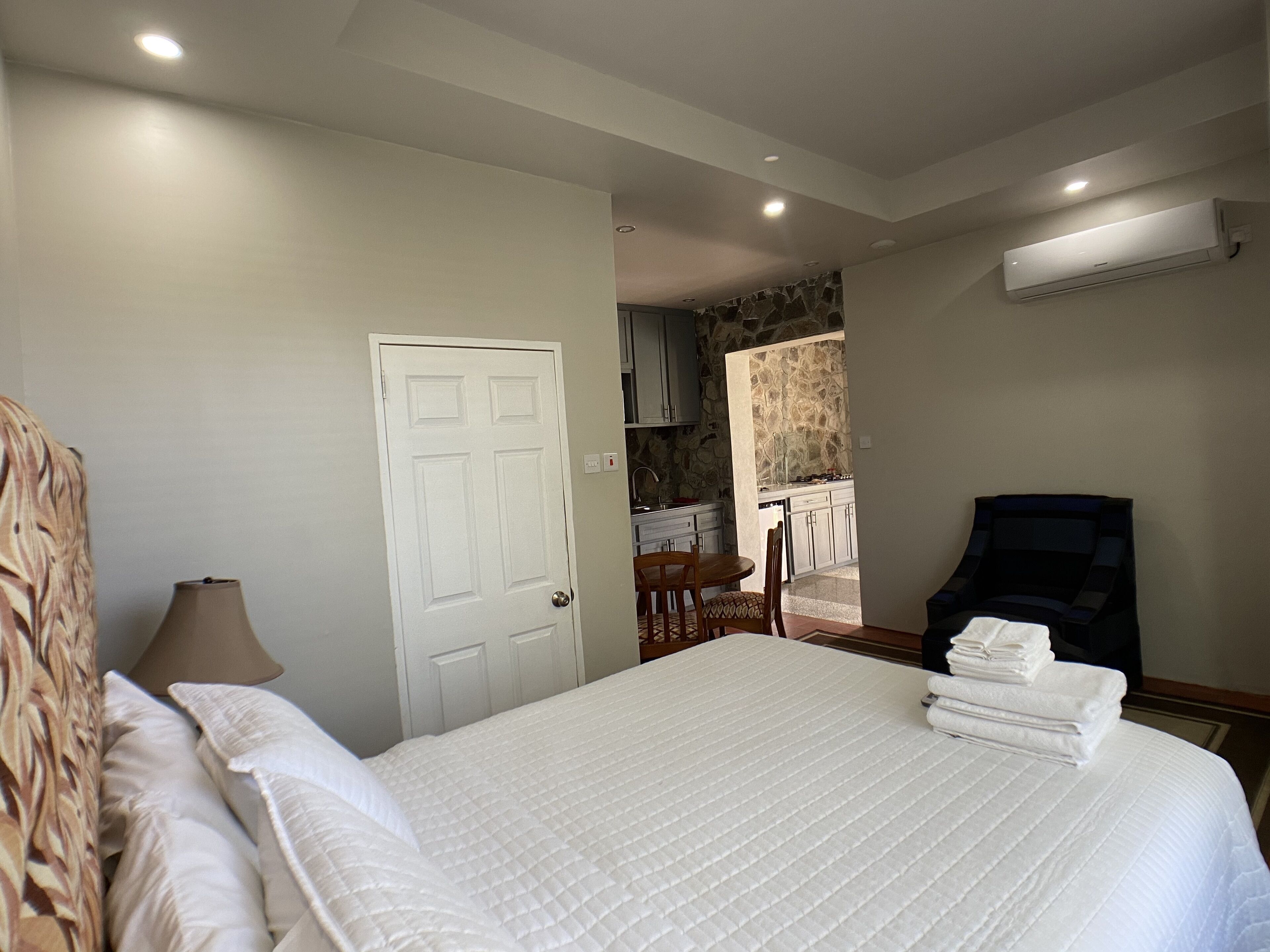 A hotel room with a bed, a chair, a desk, and a kitchenette in the background.