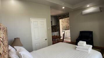 A hotel room with a bed, a chair, a desk, and a kitchenette in the background.