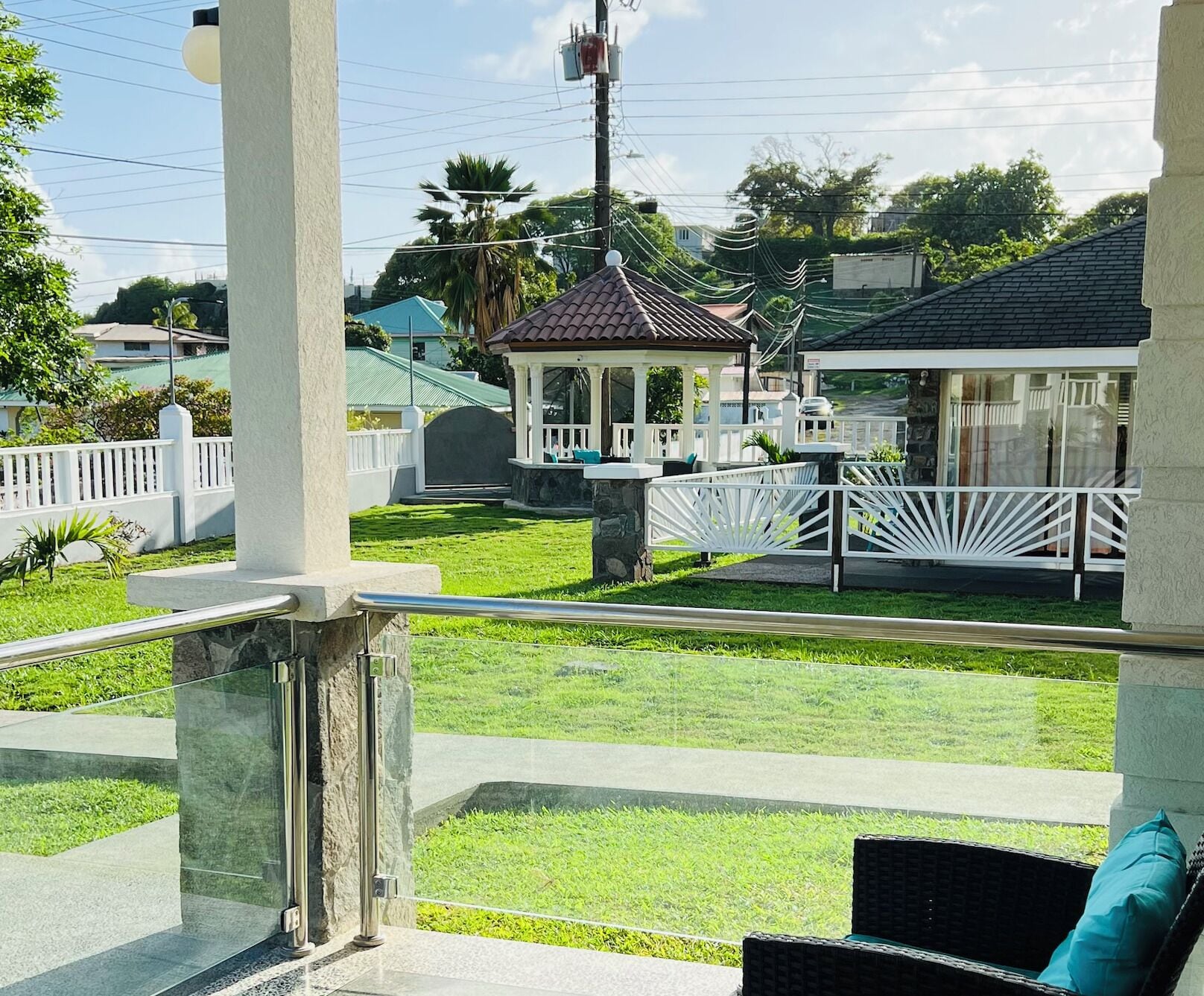 A covered outdoor seating area with a stone pillar, a white fence, and a view of a house with a tiled roof.