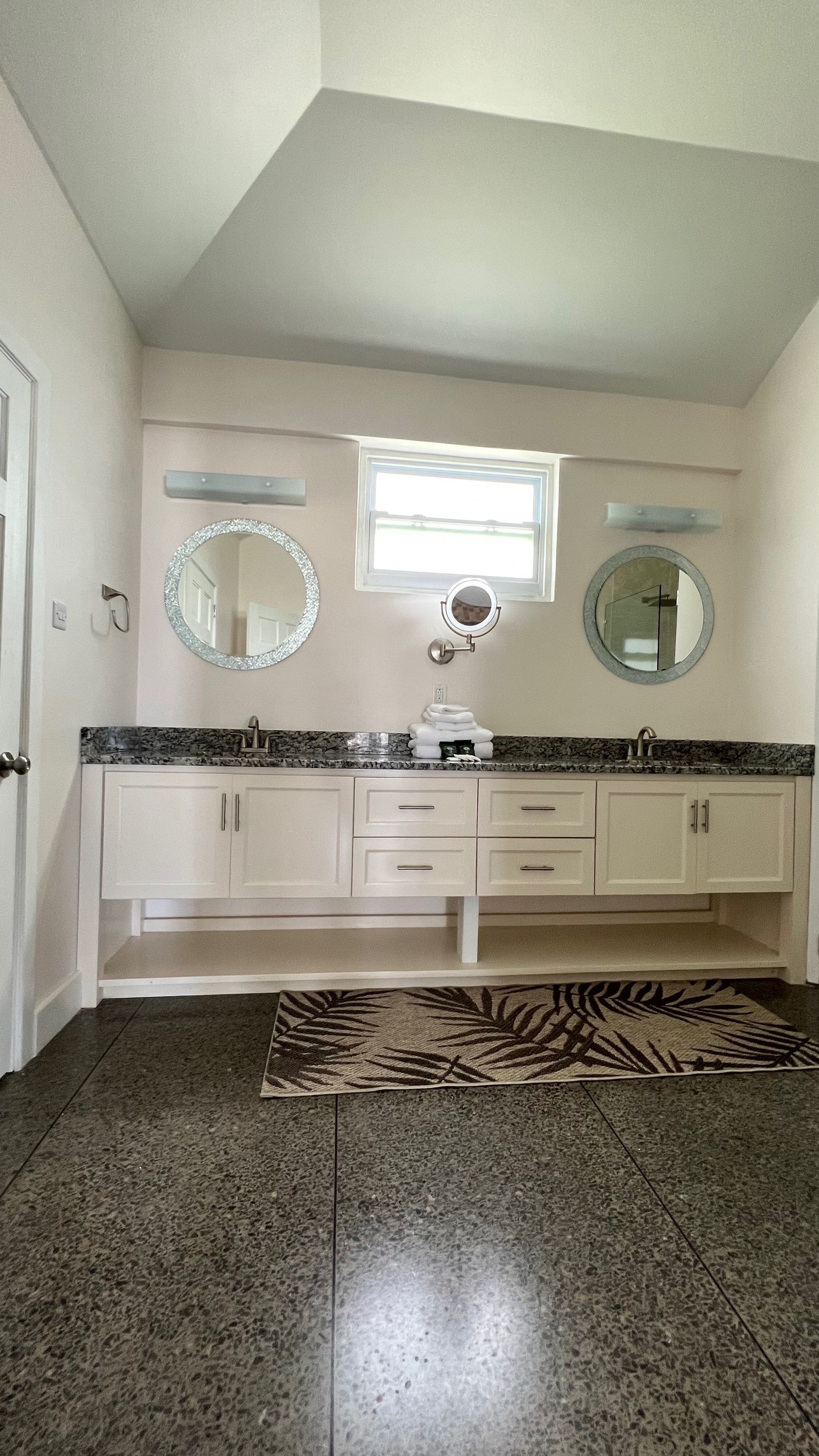 A bathroom with a granite countertop, two sinks, a vanity with drawers, a mirror, and a patterned rug on the floor.