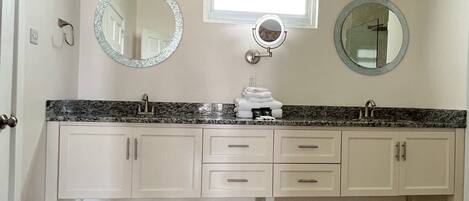 A bathroom with a granite countertop, two sinks, a vanity with drawers, a mirror, and a patterned rug on the floor.