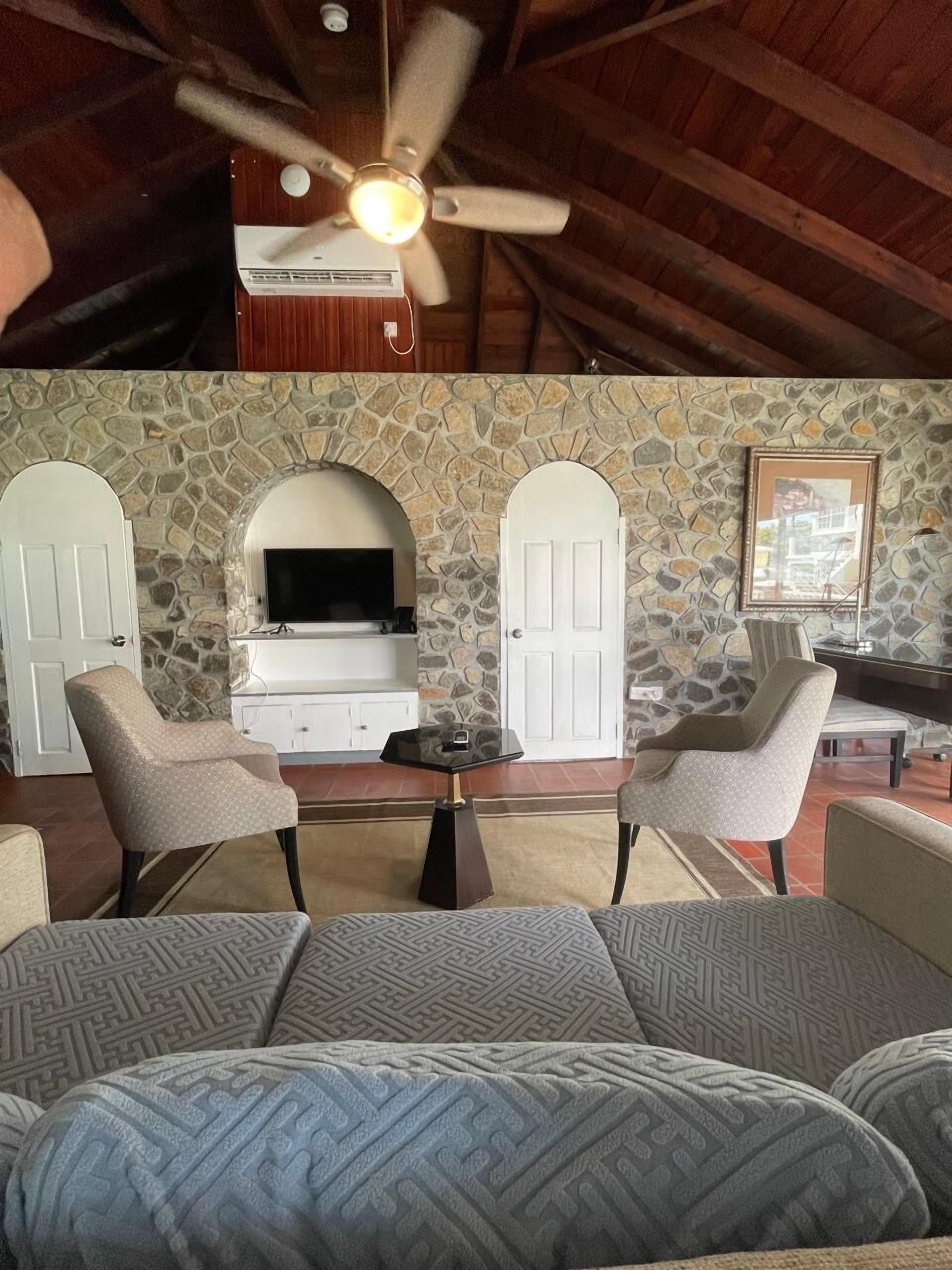 Une pièce chaleureuse avec une cheminée en pierre, deux fauteuils, un canapé, un ventilateur de plafond et un téléviseur.