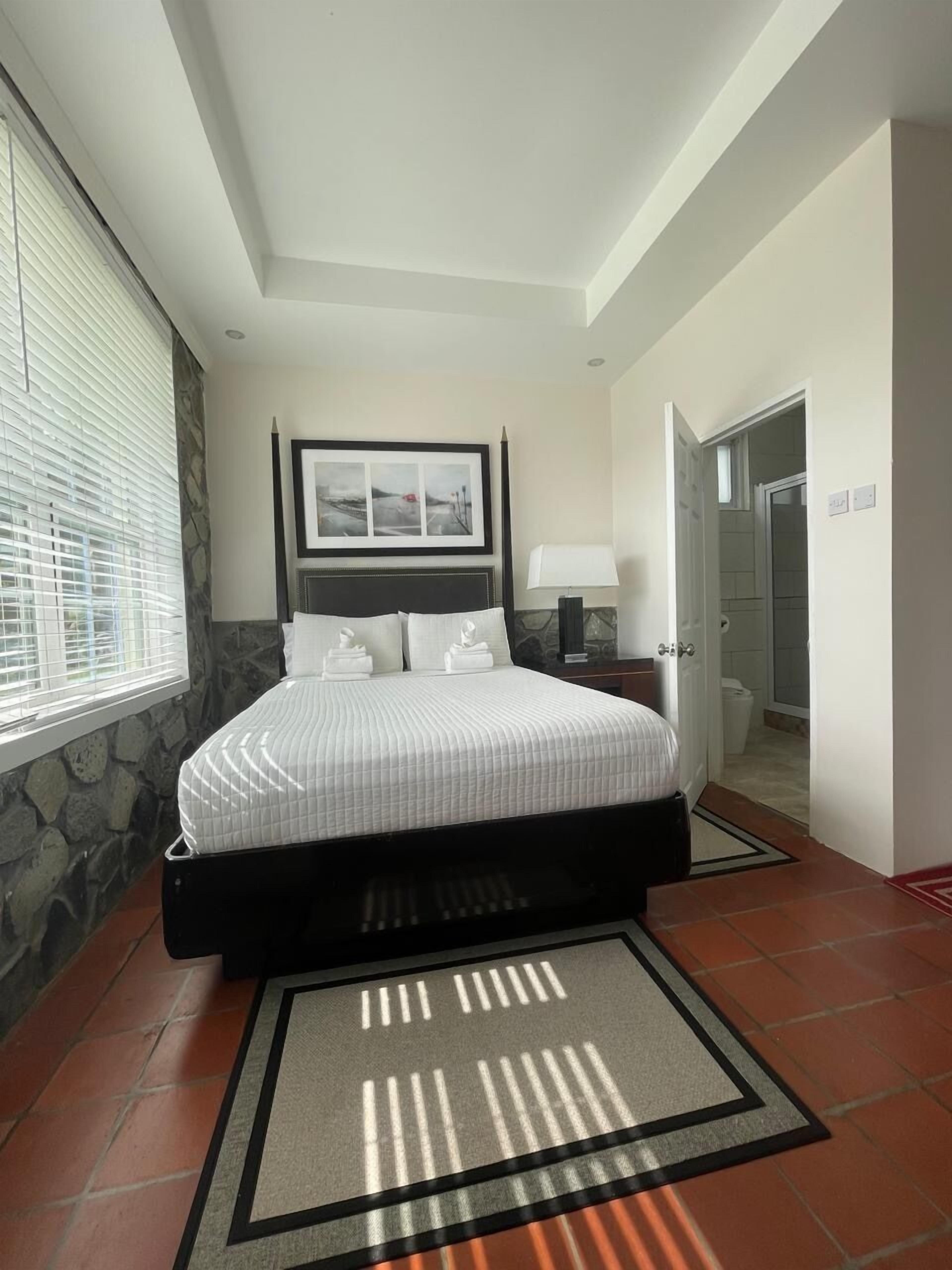 A bedroom with a bed, a nightstand, a lamp, a framed picture, and a bathroom door.