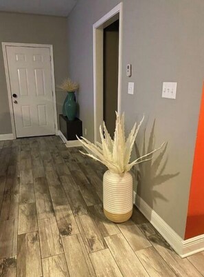 A hallway with a door at the end, featuring a tall vase with a plant and a small electrical outlet on the wall.